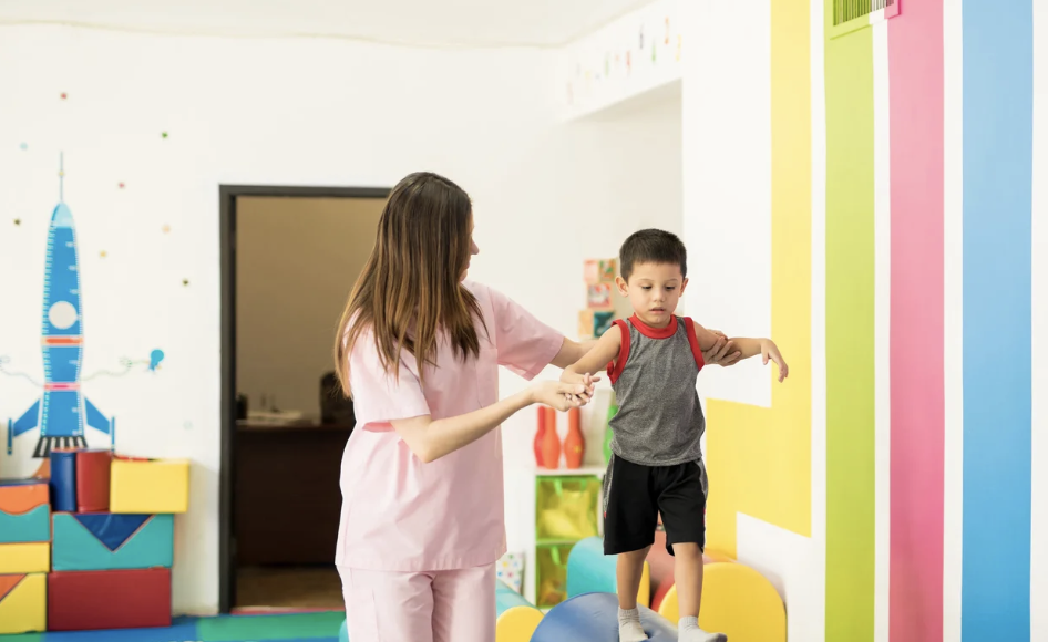 A young boy balancing on a blue exercise ball with help from a woman, in a colorful playroom.