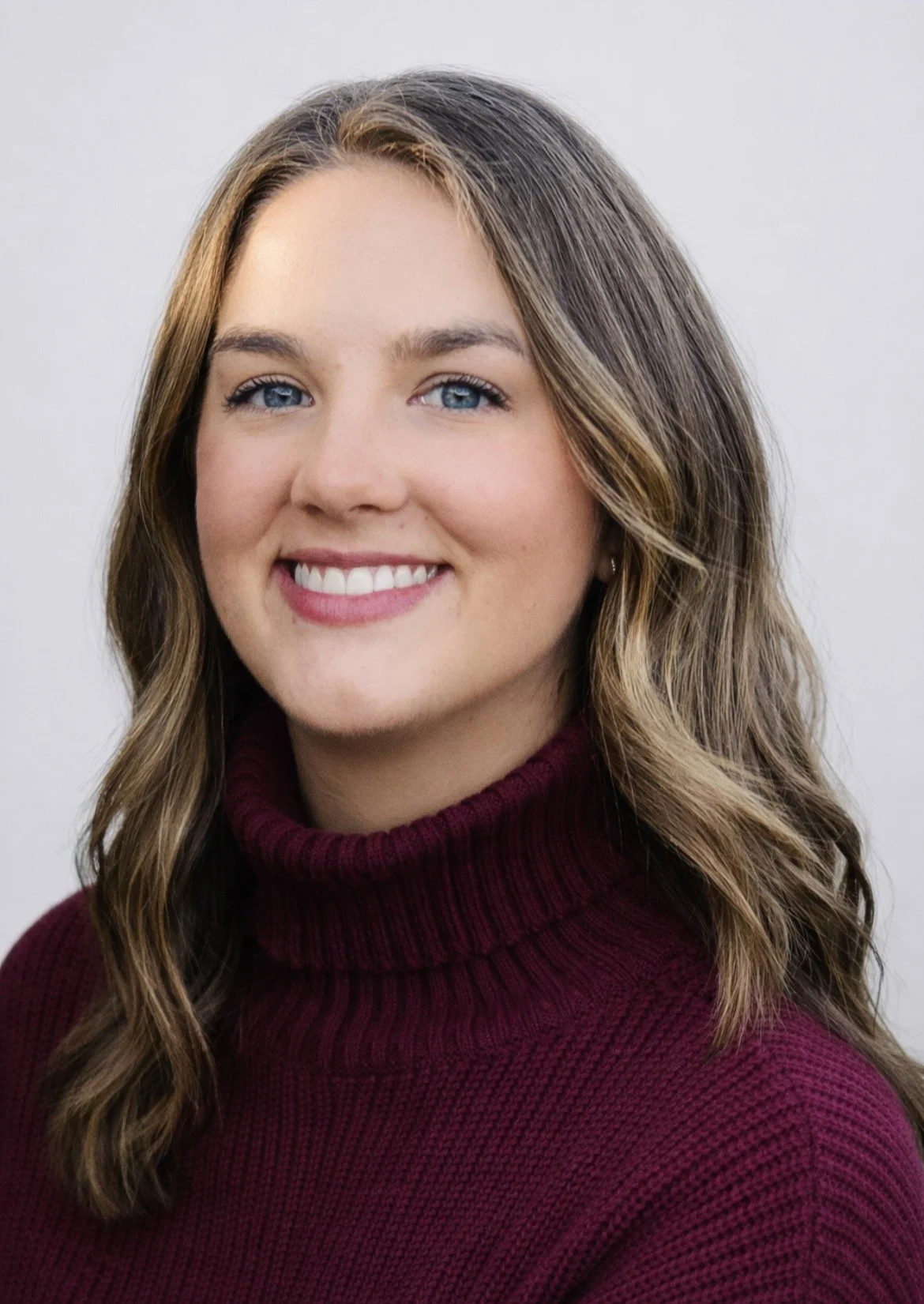 A woman with shoulder-length wavy brown hair, blue eyes, and fair skin, smiling, wearing a maroon turtleneck sweater, against a plain light gray background.