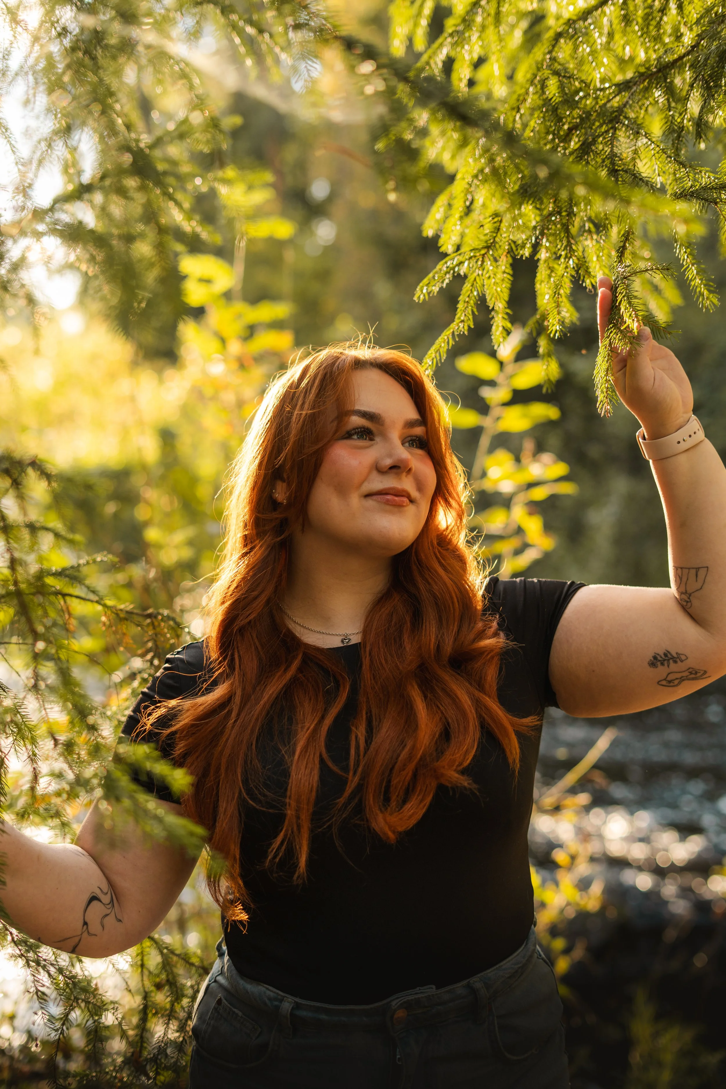 Nuori nainen, jolla pitkät punaiset hiukset ja tatuointeja, seisoo luonnossa auringon paistaessa ja koskettaa oksaa puusta.