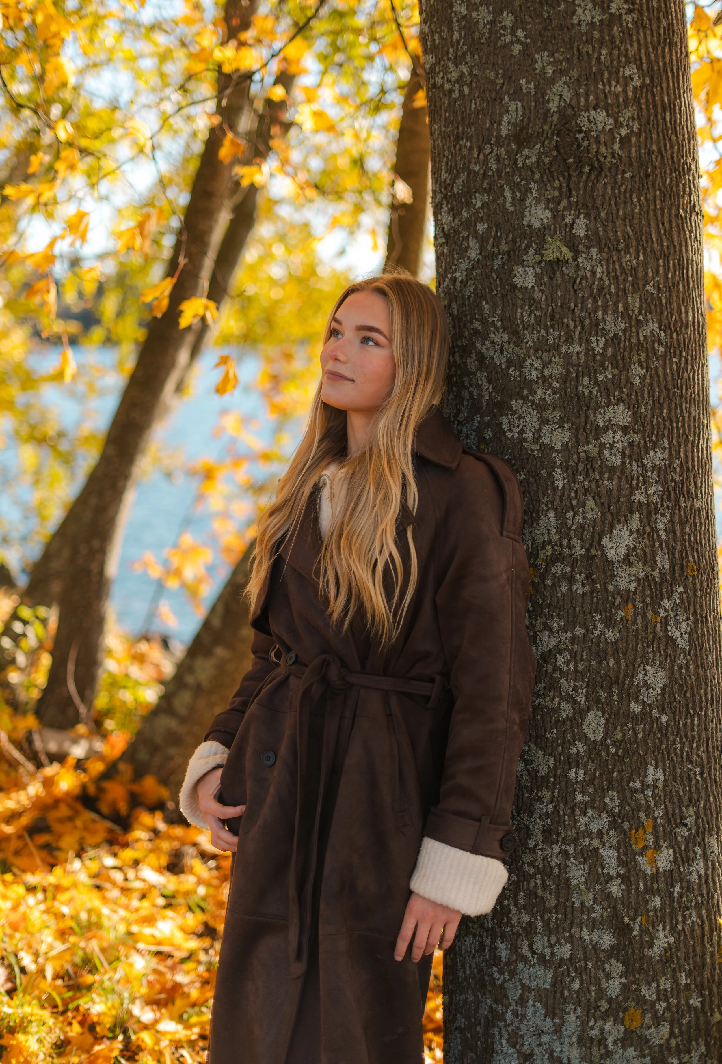 Nuori nainen nojaa puuhun syksyisessä maisemassa, taustalla lehtiä keltaisina ja oransseina.