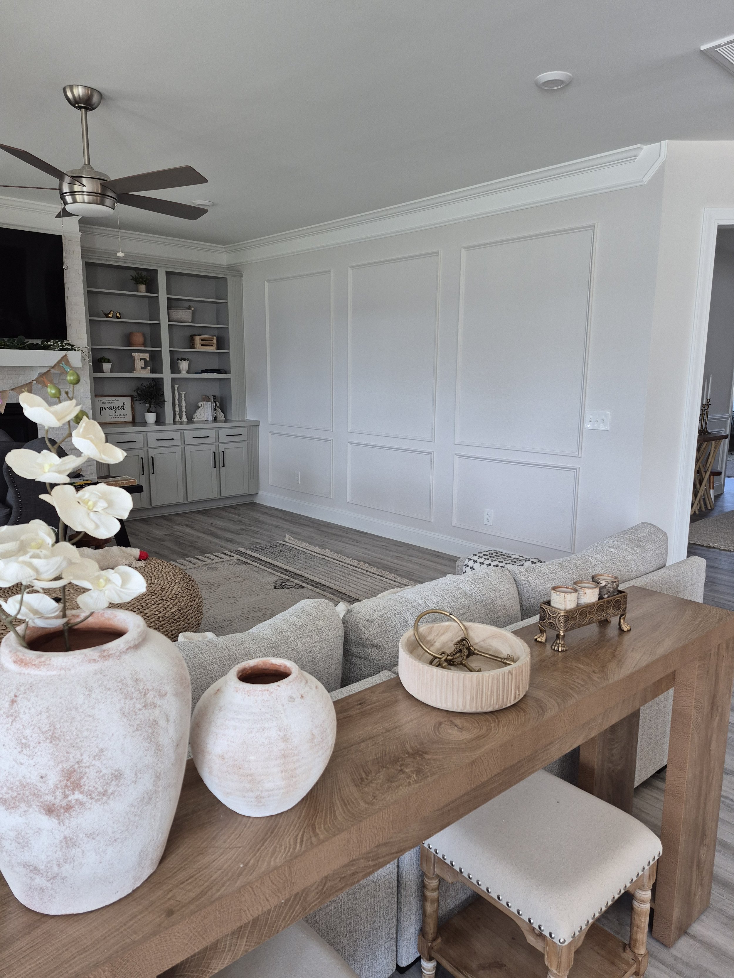 A modern living room with a ceiling fan, built-in gray shelving, white paneled walls, a beige sofa, and decor including vases, candles, and a small chair.