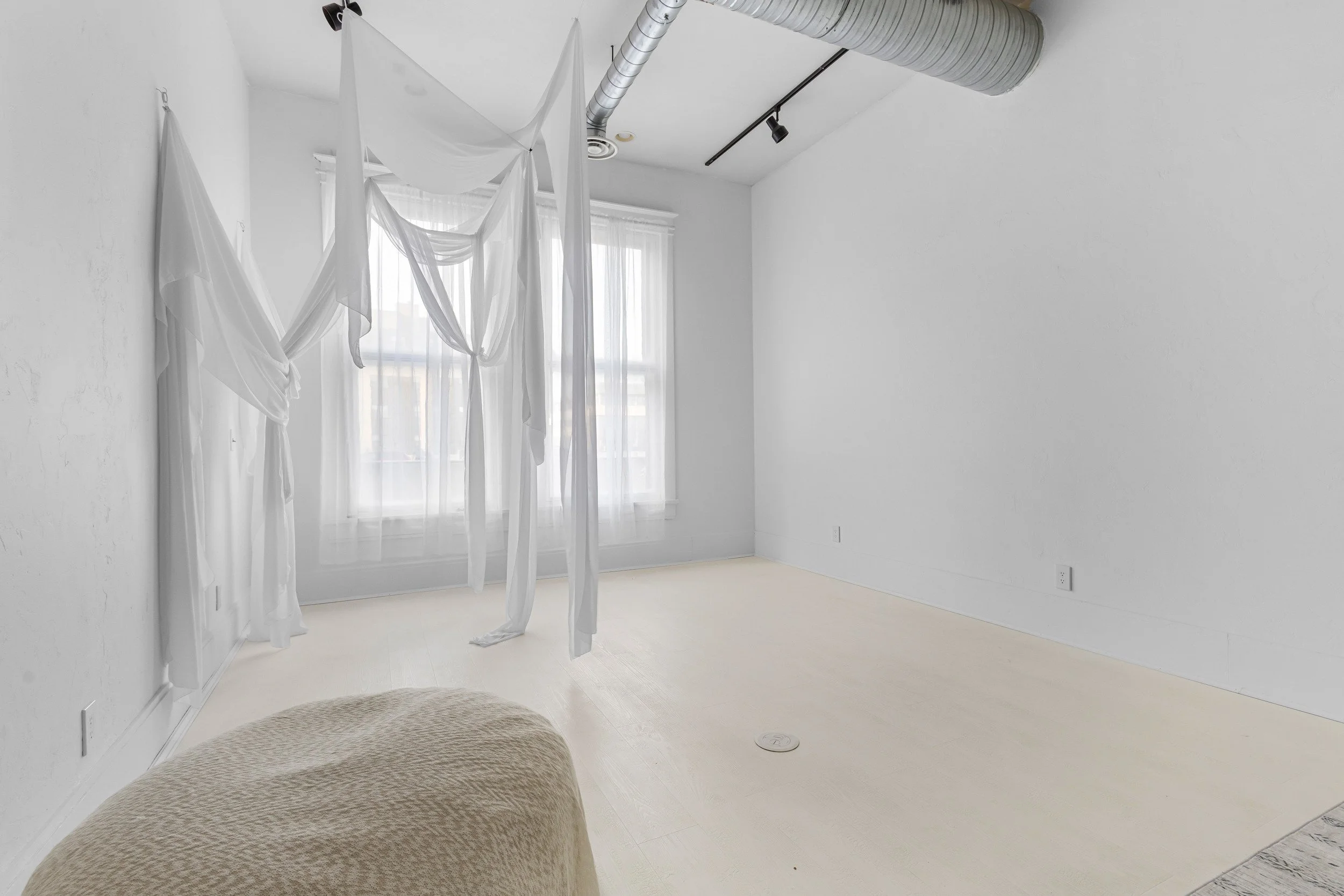 Empty room with white walls, light-colored flooring, large windows with sheer curtains, and hanging white fabric or sheets from the ceiling.