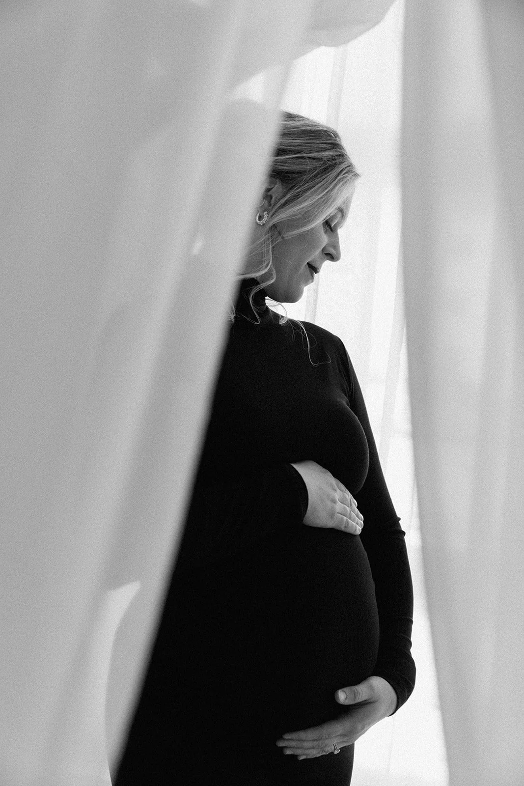 Black and white photograph of a pregnant woman standing indoors, her hand resting on her belly, with curtains and a window in the background.