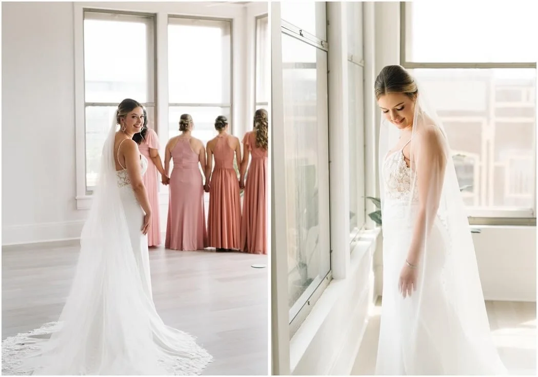 A bride in a white wedding gown with a lace bodice and veil stands beside a window, smiling down. Behind her, a group of bridesmaids in matching pink dresses hold hands and look at her, standing near large windows in a bright room.