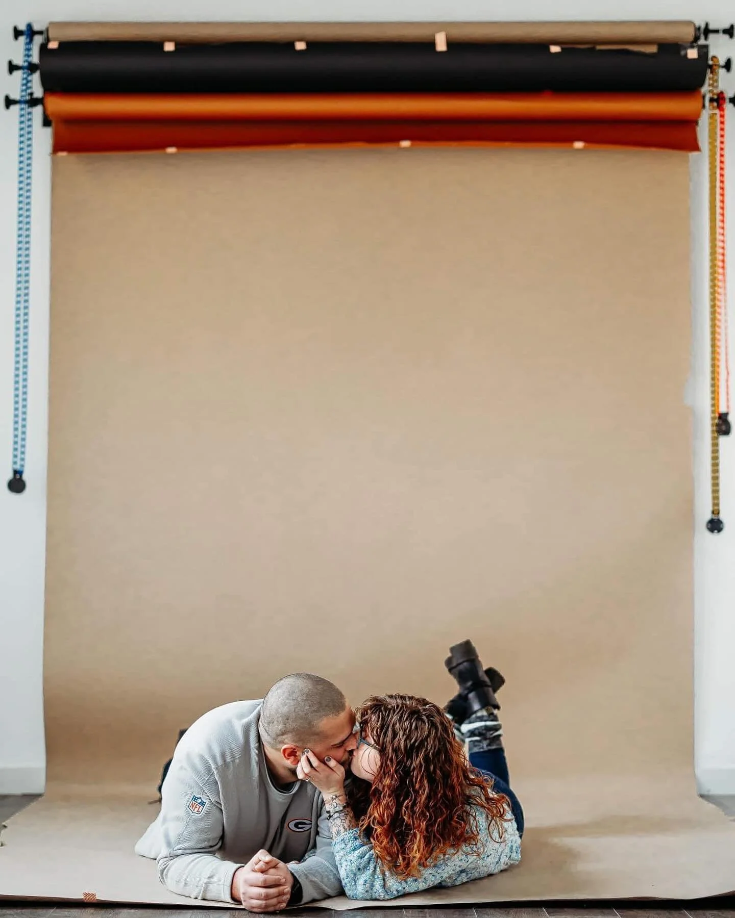 A couple lies on their stomachs on a brown paper backdrop, sharing a kiss. The woman has curly red hair and is wearing a sweater, while the man has short hair and is wearing a gray NFL jersey.