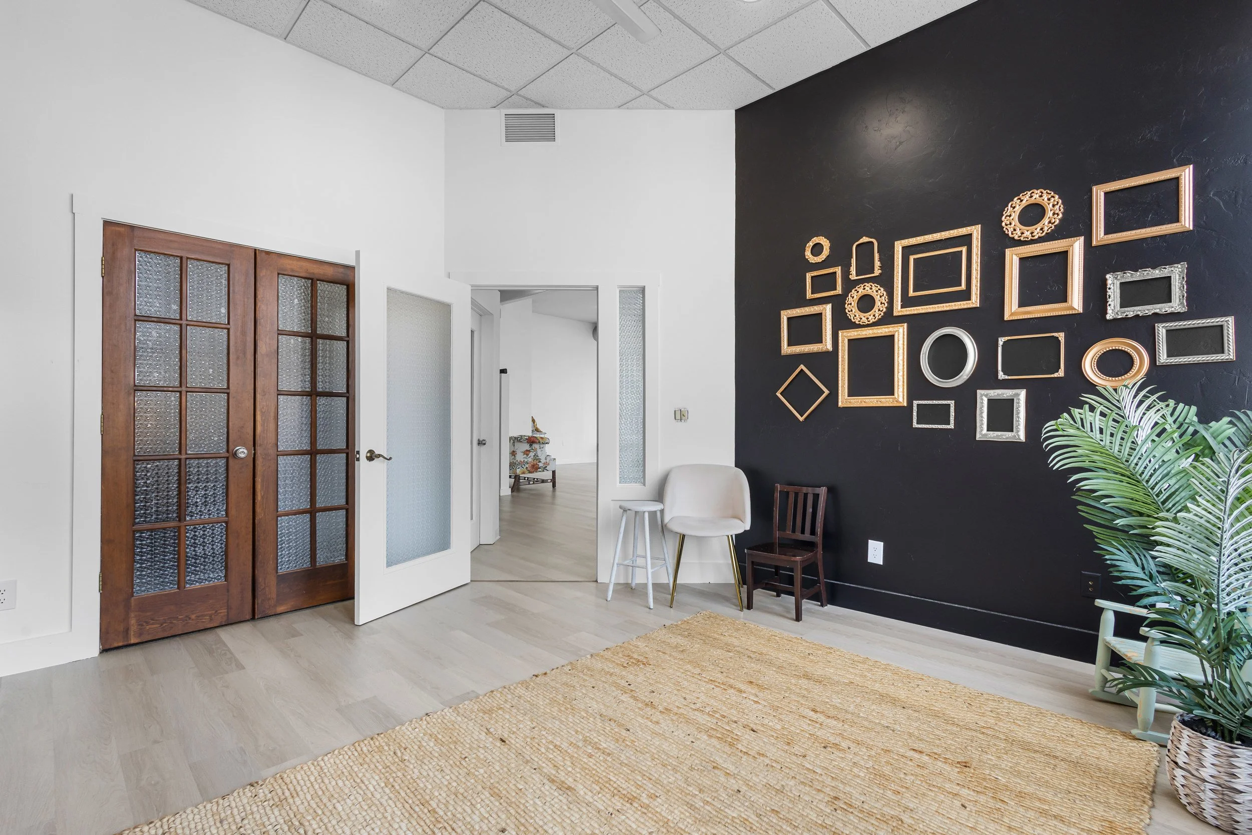 A contemporary living room corner with a black accent wall decorated with an array of framed mirrors and art, beige armchair, small white stool, plant in a woven basket, and wooden doors with glass panels on light wood flooring.