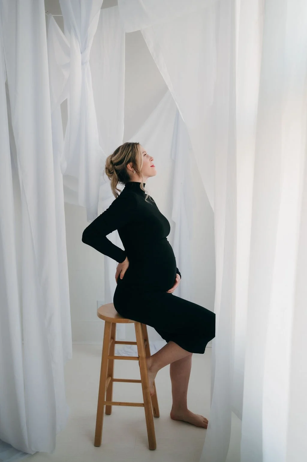 Pregnant woman in black dress sitting on a stool, with her eyes closed and hand on her belly, surrounded by white flowing curtains.