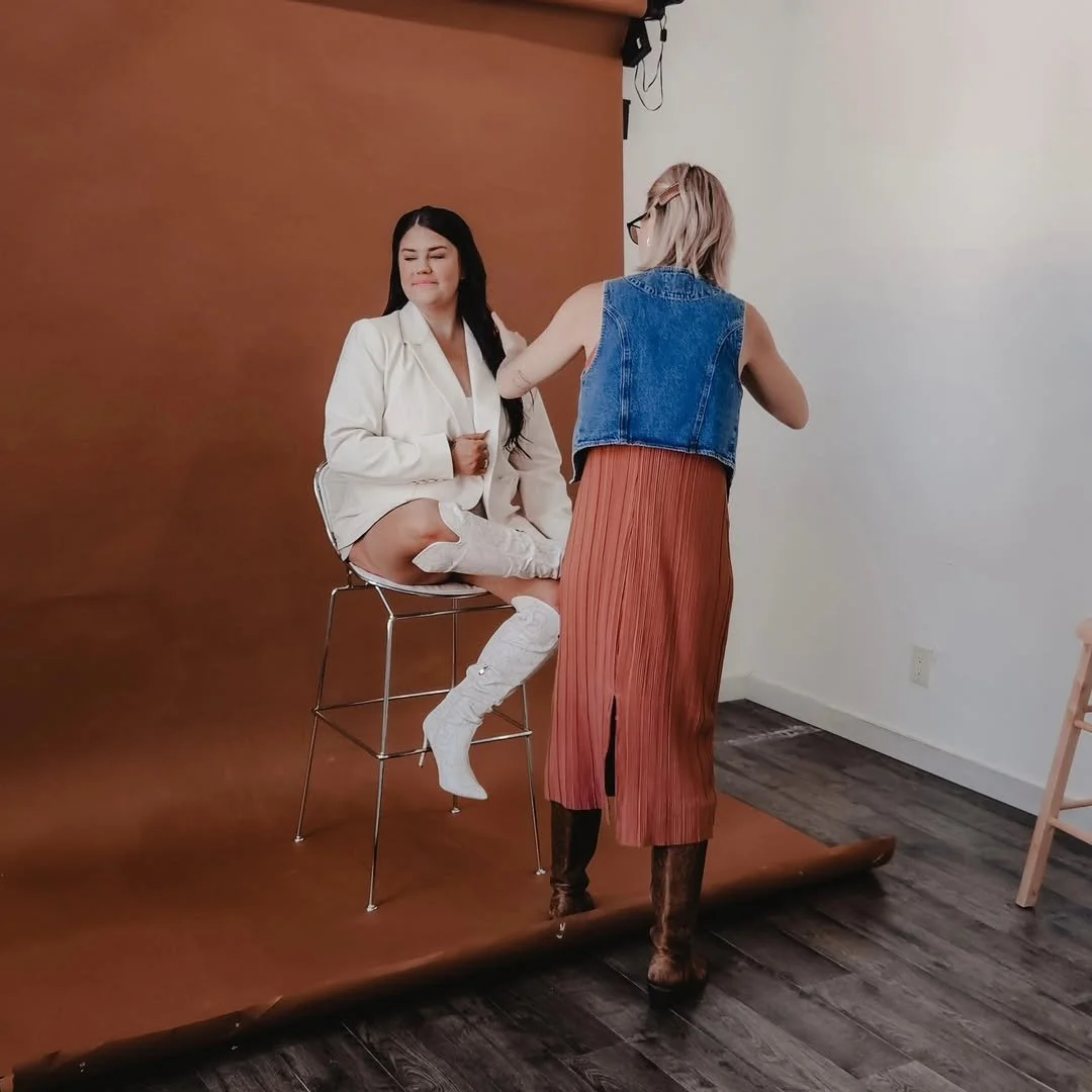 A woman with long dark hair sits on a high stool with her eyes closed, dressed in a white blazer and thigh-high white boots. A stylist with blonde hair, glasses, a denim vest, and a rust-colored pleated skirt styles her. They are in front of a brown