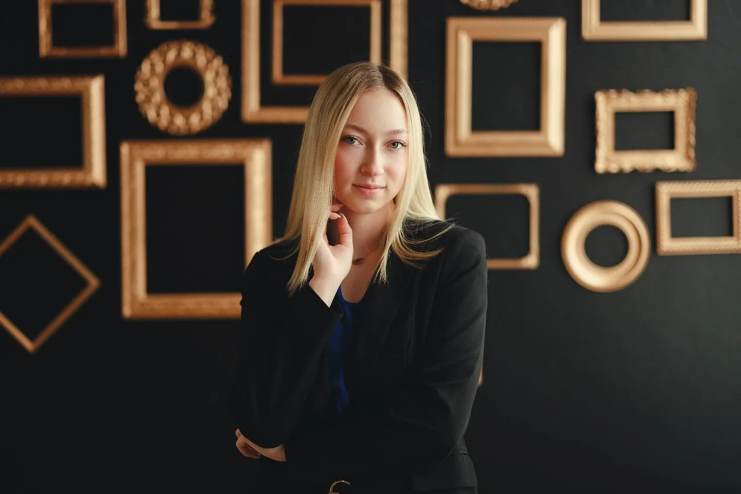 A young woman with blonde hair dressed in a black blazer and blue top, standing in front of a black wall decorated with empty gold picture frames, looking at the camera with her hand near her face.