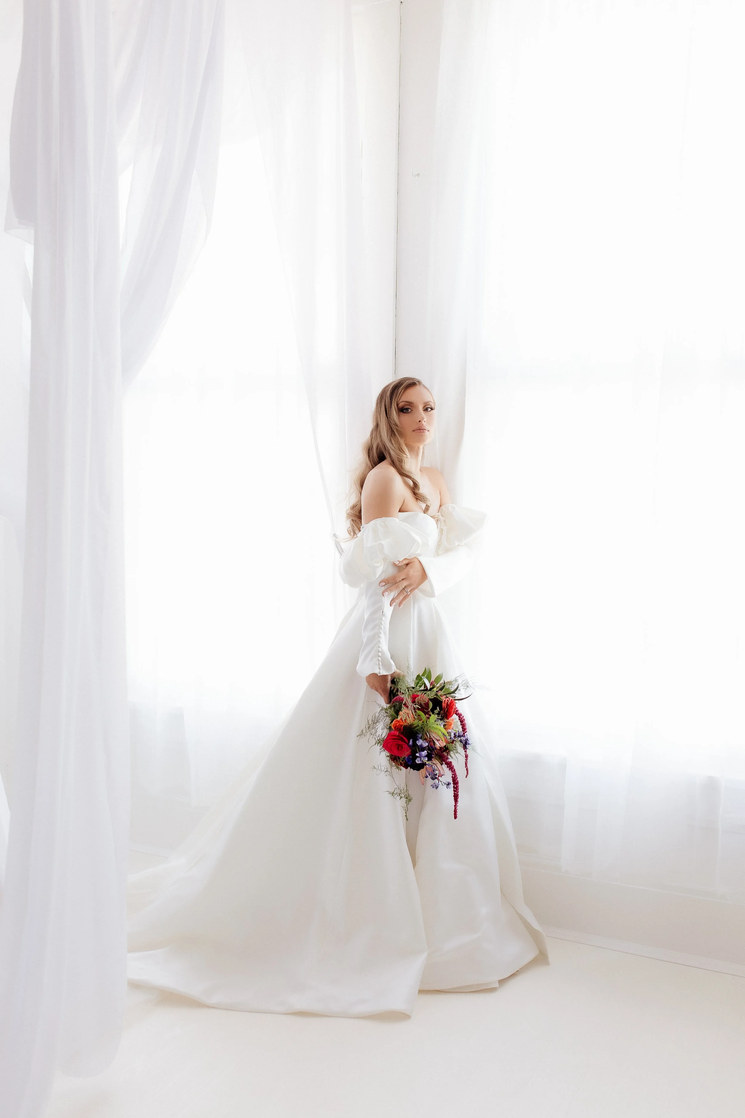 Bride in a white wedding gown holding a colorful bouquet, standing near large windows with white curtains, soft natural light illuminating the scene.