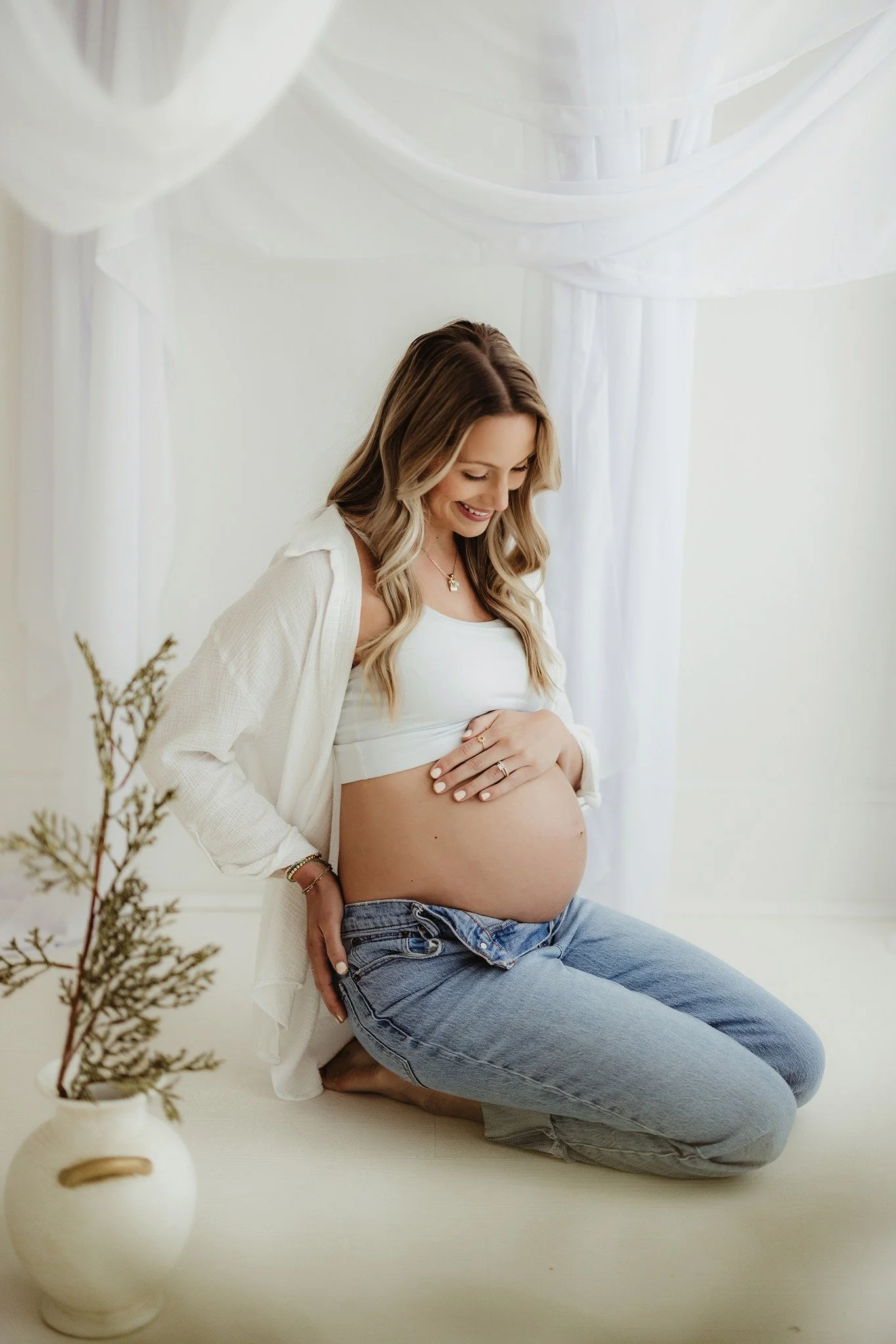 Pregnant woman kneeling on the floor, smiling and holding her stomach, in a bright, white room with sheer curtains and a decorative vase with branches in the foreground.