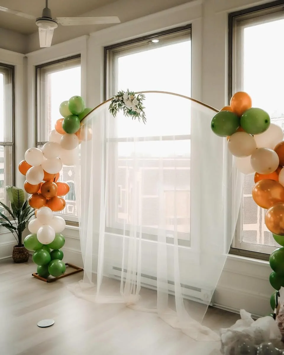 Balloon arch with green, white, and orange balloons arranged in clusters on each side of a sheer curtain backdrop, decorated with a small floral arrangement at the top center, set up indoors near large windows.