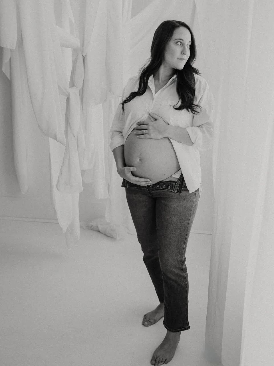 Pregnant woman with long dark hair standing barefoot in a room with hanging white fabric, lifting her shirt with one hand and resting the other on her belly, looking to the side.