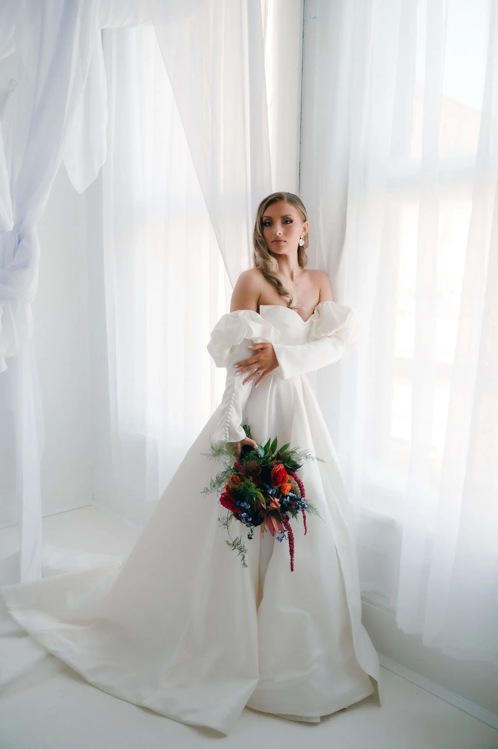 Bridal woman in white wedding dress holding a colorful bouquet of flowers, standing by a window with white curtains.