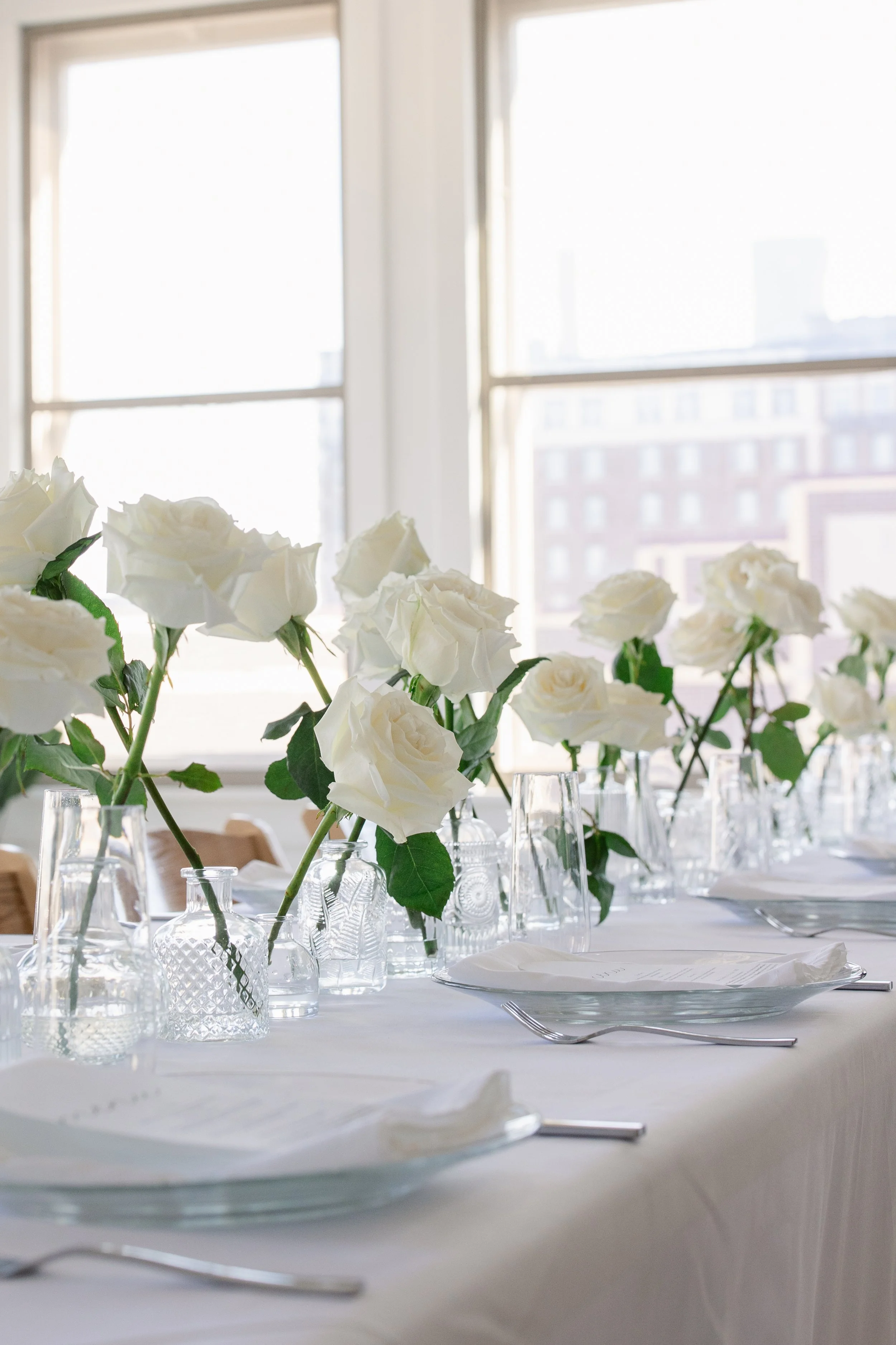 Elegant table setting with white roses in glass vases, wine glasses, plates, napkins, and silverware near large windows with city views.