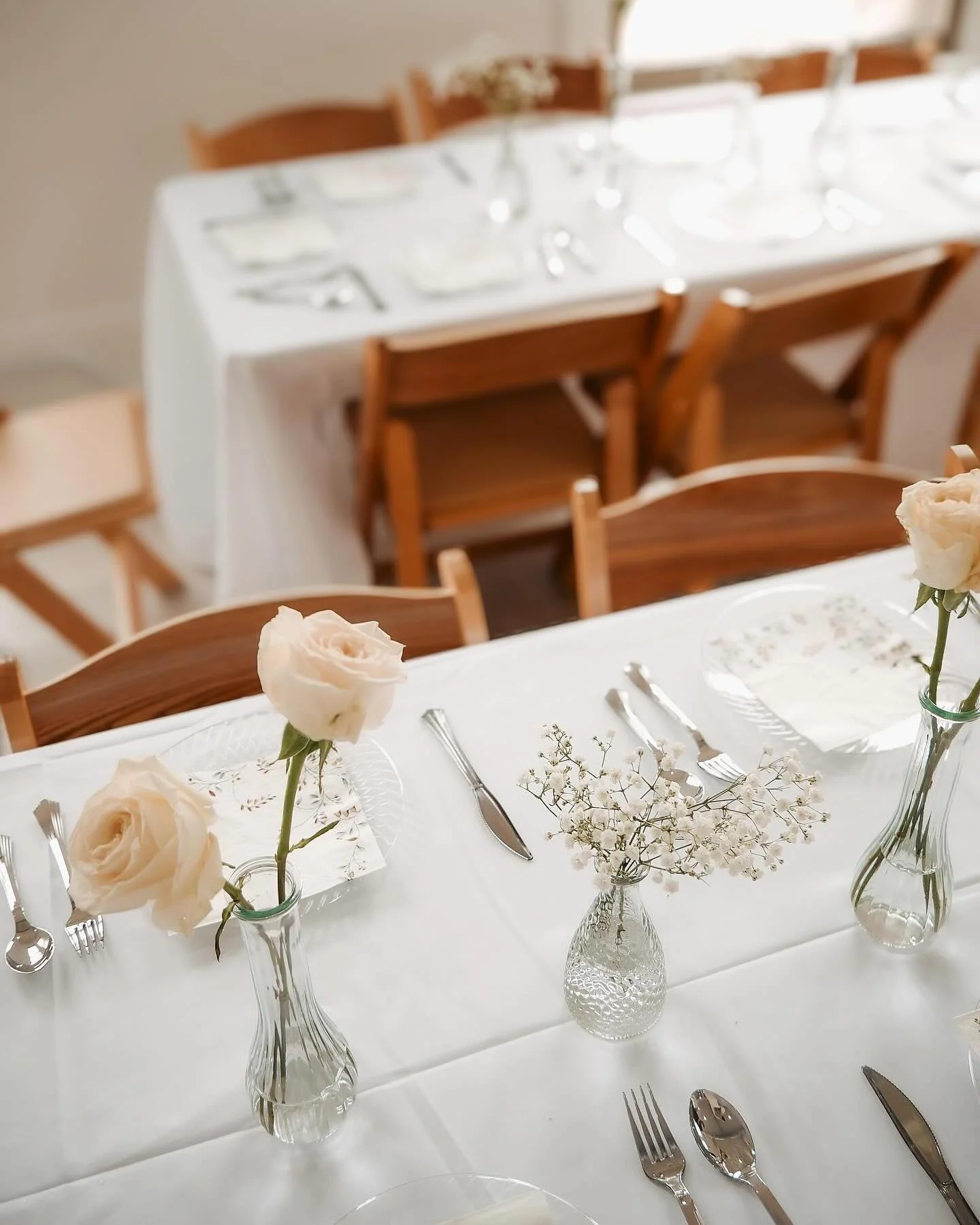 A formal dining table decorated with white flowers in glass vases, set with silverware, napkins, and plates, with a cozy dining area in the background.