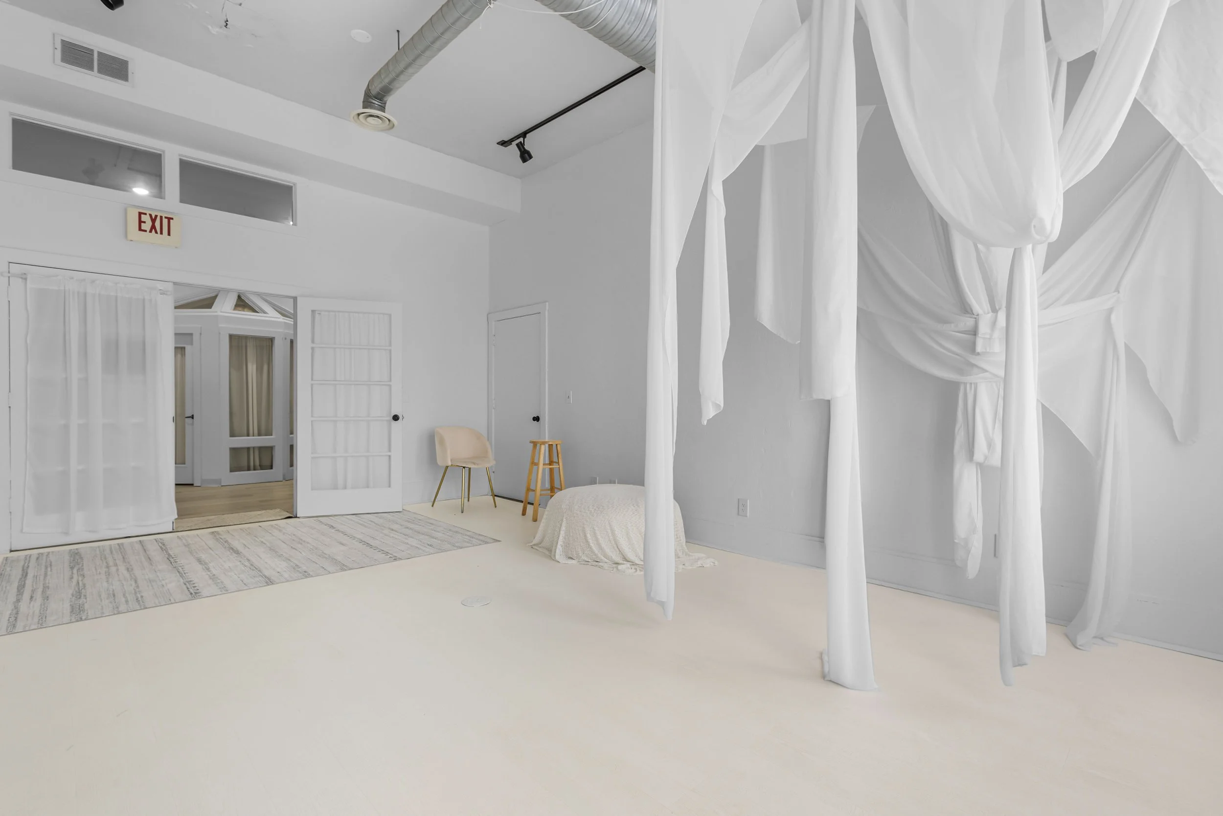 Large, bright, minimalist room with white walls and floor, featuring a four-poster bed with white curtains, a beige chair, a small wooden stool, a patterned rug, and an exit door with glass panels.
