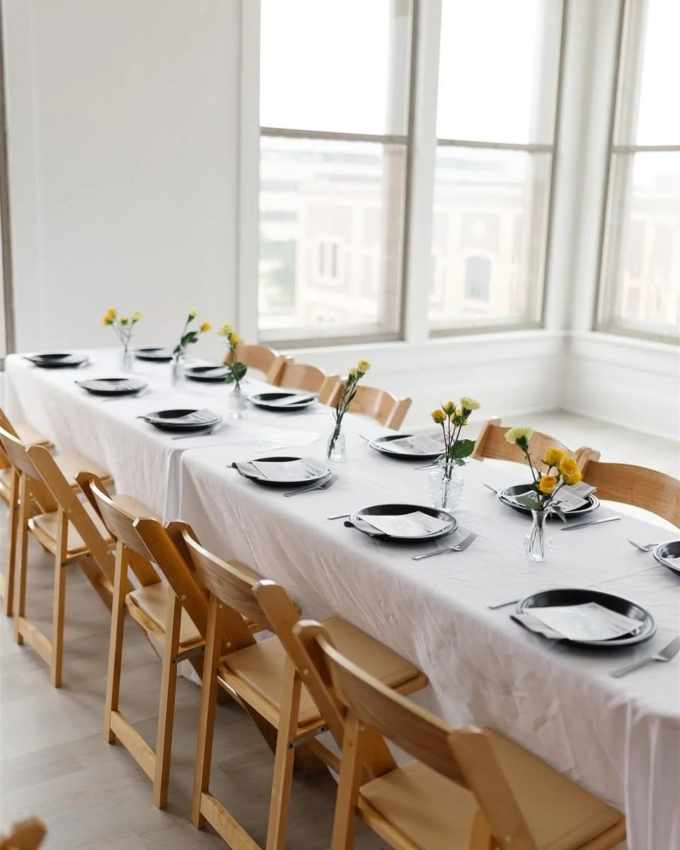 Long dining table set for a meal with black plates, silverware, and small vases with yellow flowers, in a room with large windows overlooking a cityscape.