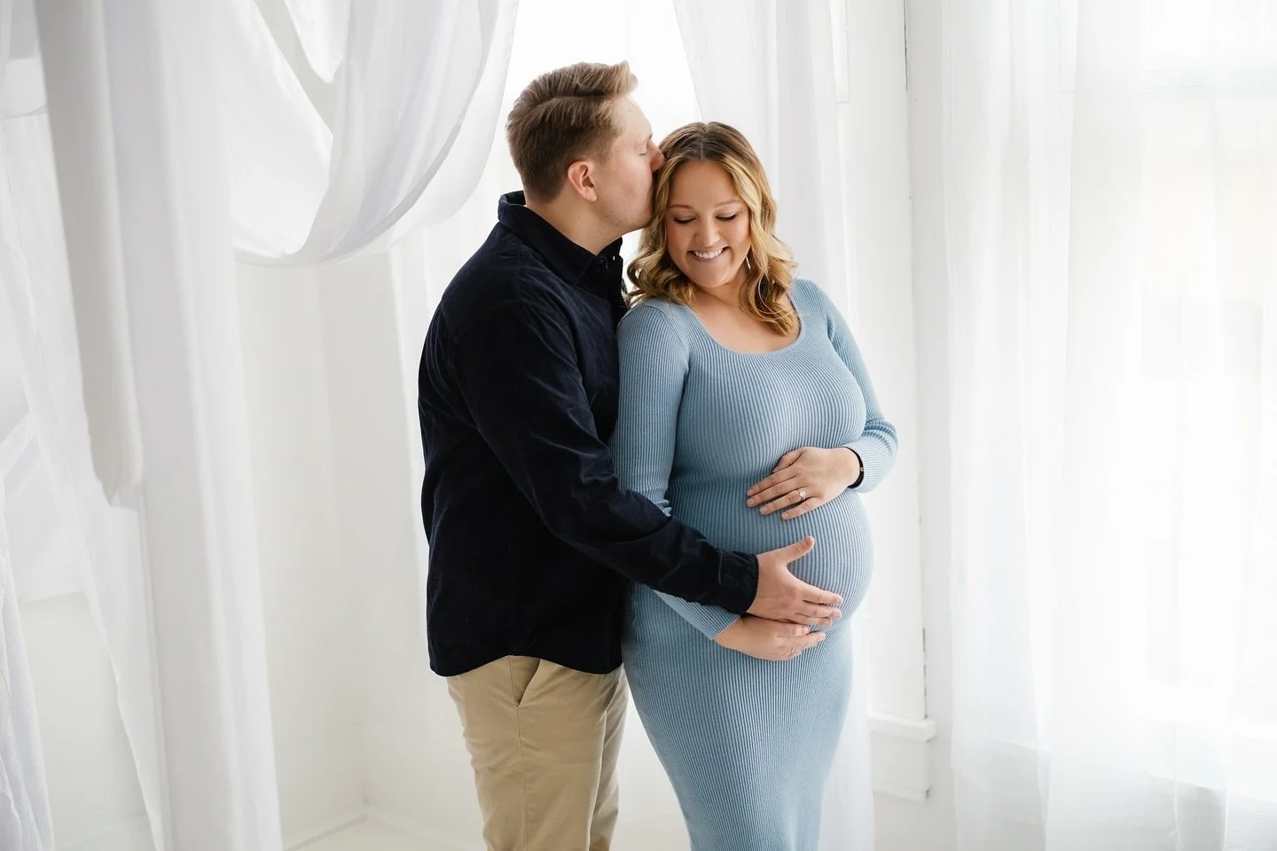 A man kissing a pregnant woman on the forehead, smiling and holding her baby bump in a bright, white room with sheer curtains.