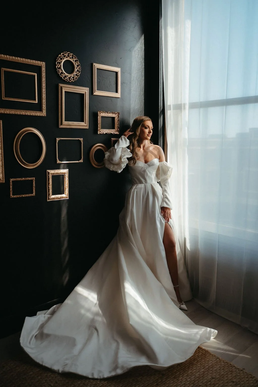 A woman in a white off-shoulder wedding gown standing near a window with sheer curtains, with a dark wall decorated with empty picture frames behind her.
