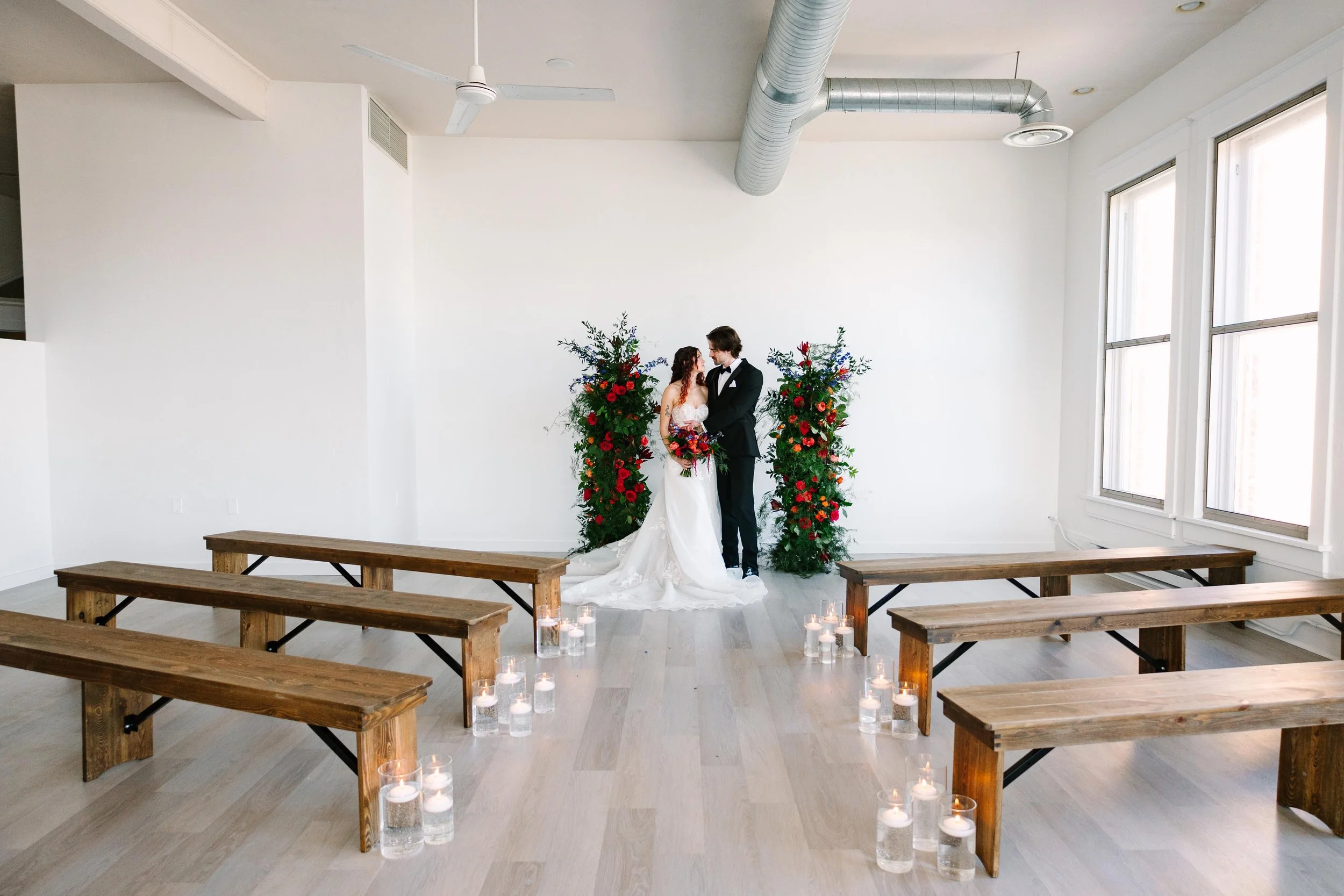 A bride and groom standing together on a small indoor wedding stage with floral arrangements behind them and lit candles on the floor.