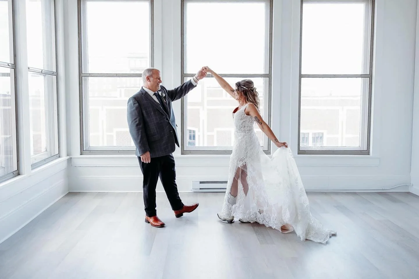 A bride and an older man, possibly her father, dancing together in a bright, minimalist room with large windows. The father is holding the bride's hand as she twirls.