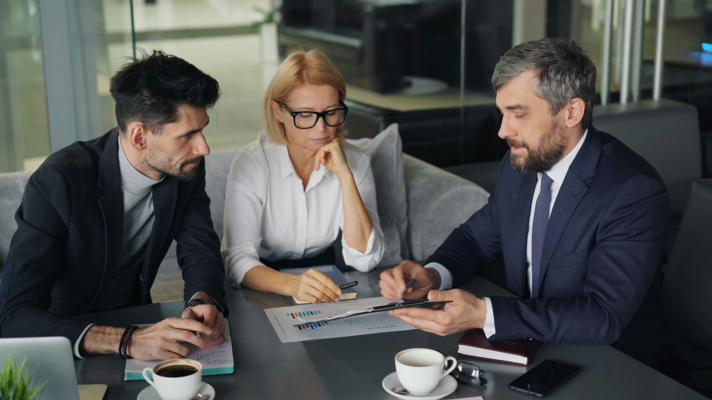 Three professionals in business attire engaged in a serious discussion at a conference table, reviewing documents and notes, in a modern office setting.