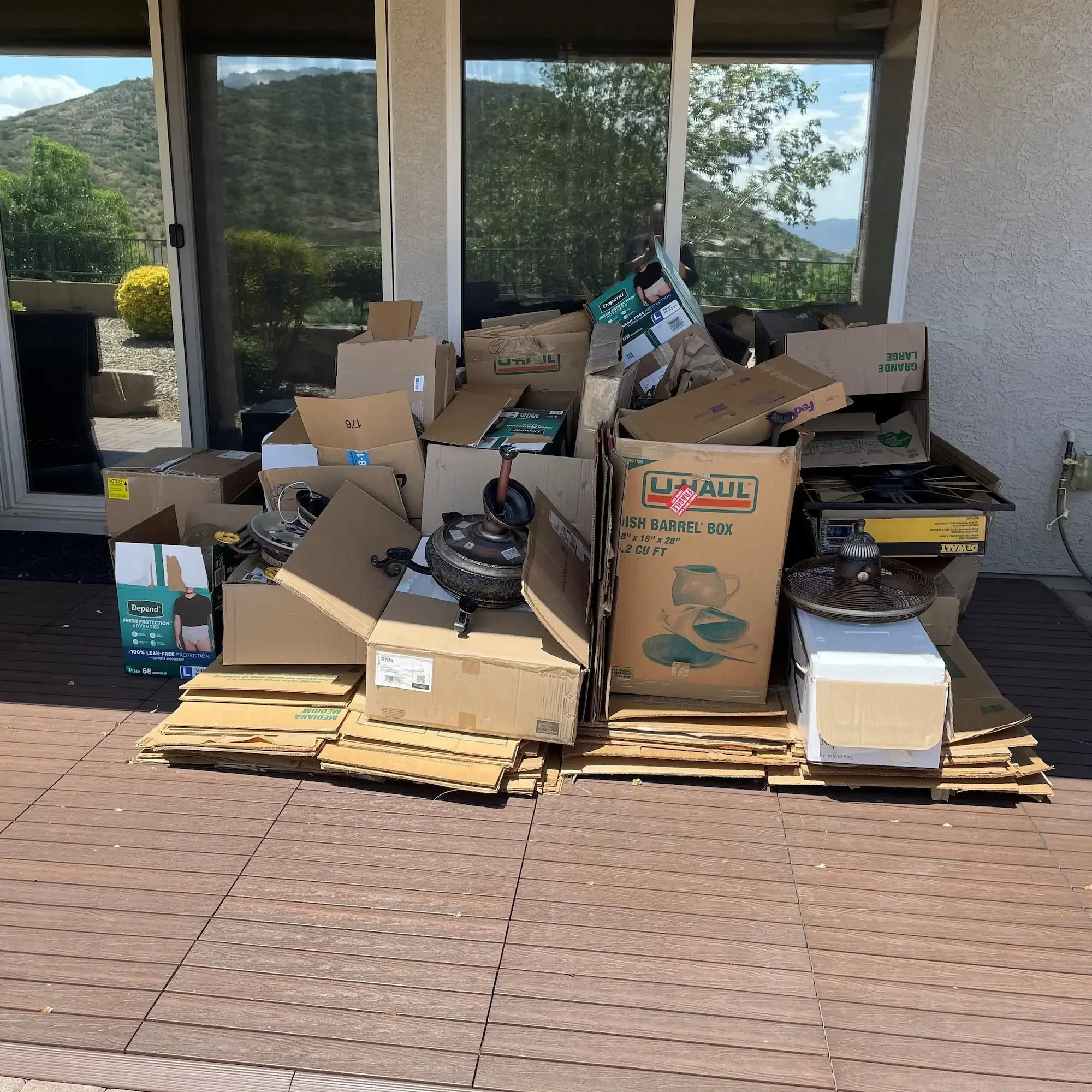 Stack of moving boxes on a wooden deck with some household items, next to a sliding glass door with a mountain view in the background.
