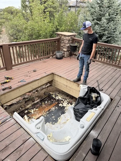 A person wearing a mask and gloves is standing on a wooden deck, observing an empty hot tub that is partially demolished. Tools are scattered on the deck, and there are trees in the background.