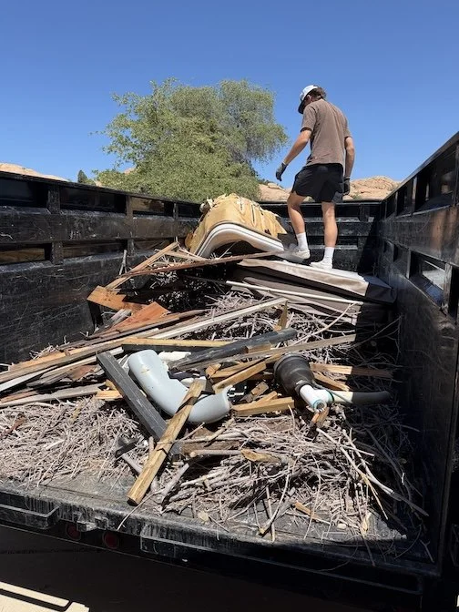 A person is standing inside a dumpster filled with debris like wood, plastic pipes, and other trash, under a clear blue sky, with a tree and rocks in the background.