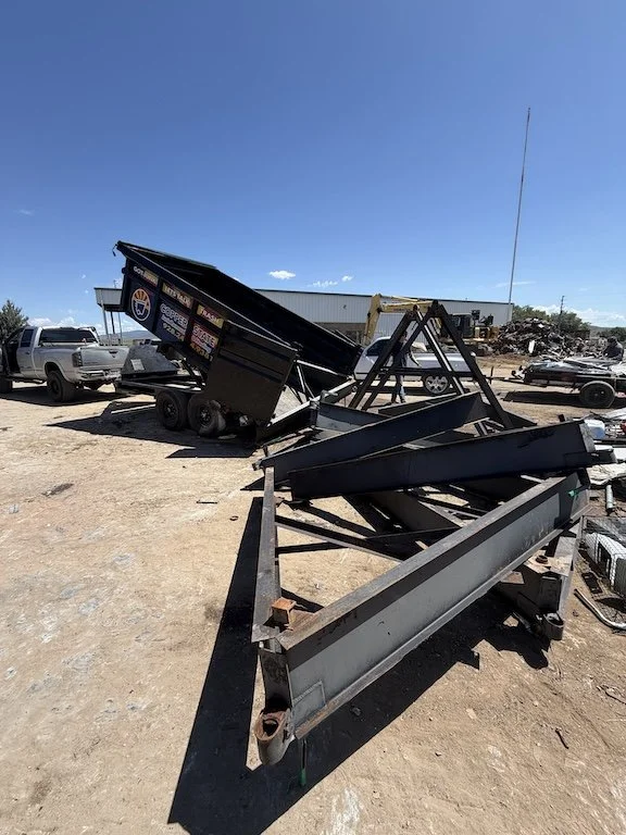Stacked trailer frames in an outdoor lot with cars and a building in the background under a clear blue sky.