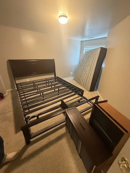 Disassembled bed frame and a separate mattress leaning against the wall in a bedroom with beige walls and a window, with a small wooden nightstand nearby.