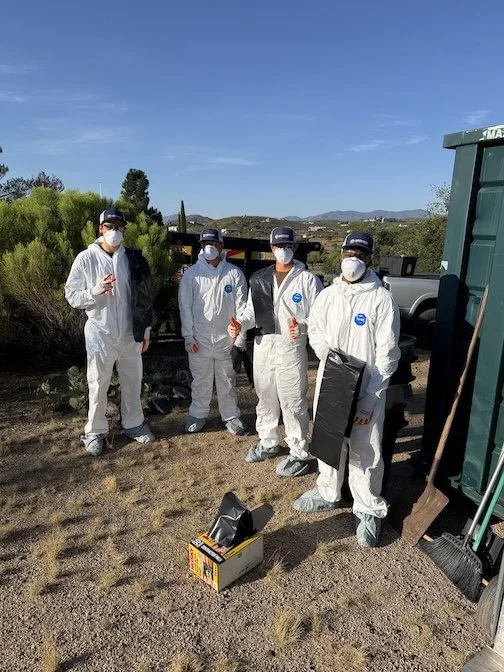 Four people wearing white protective suits, face masks, and caps standing outdoors near a green dumpster and a tree. They are holding orange tools and a black object, with a box on the ground in front of them. The background features a clear blue sky and some distant hills.