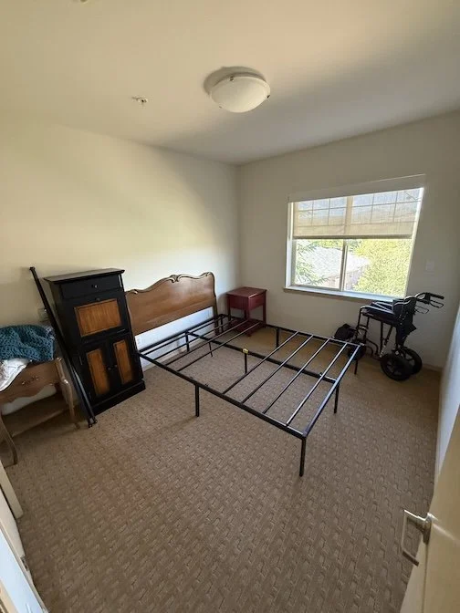 Empty bedroom with a metal bed frame, wooden headboard, small red nightstand, black bicycle, window with blinds, and beige carpet.