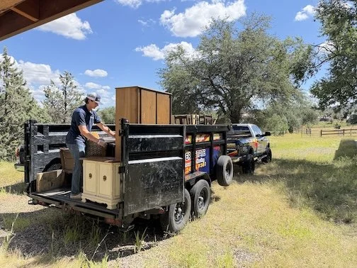 A man loading furniture onto a black pickup truck with a flatbed, parked on a grassy area near trees and a fence under a blue sky with scattered clouds.