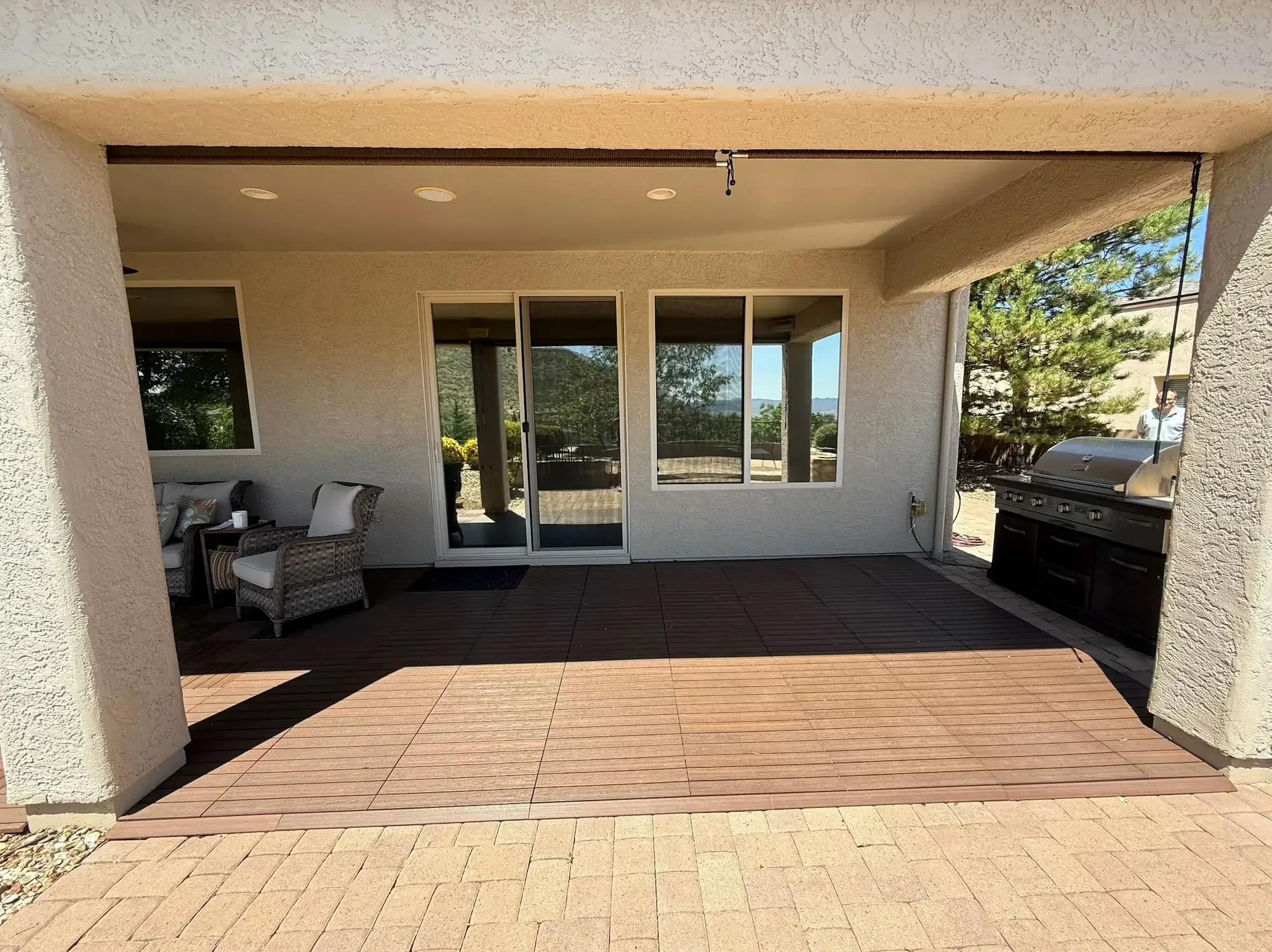 Covered outdoor patio with wicker chairs and a grill, sliding glass door, and windows, overlooking a backyard with trees and a patio area.