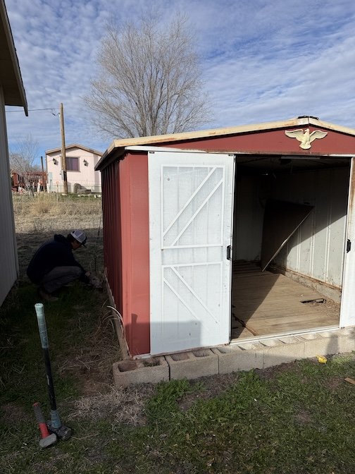 A red and white shed with open doors, inside there is a broom leaning against the wall, in a backyard with a person working with a tool near the shed, a large tree, and other houses in the background.