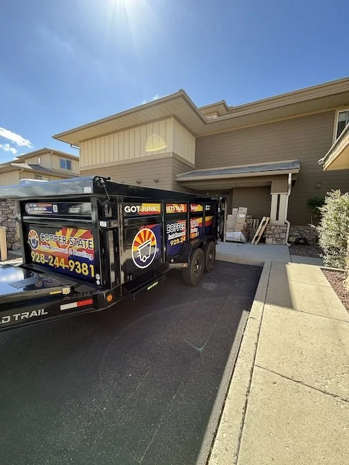 A black trailer parked on a driveway next to a beige house with a porch, with a clear sunny sky overhead.