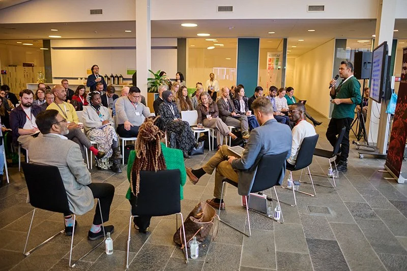 A photograph of arguably the first Common Ground session in Oslo, shown with a group of diverse people sitting in a circle on metal and plastic chairs in a brightly lit room.