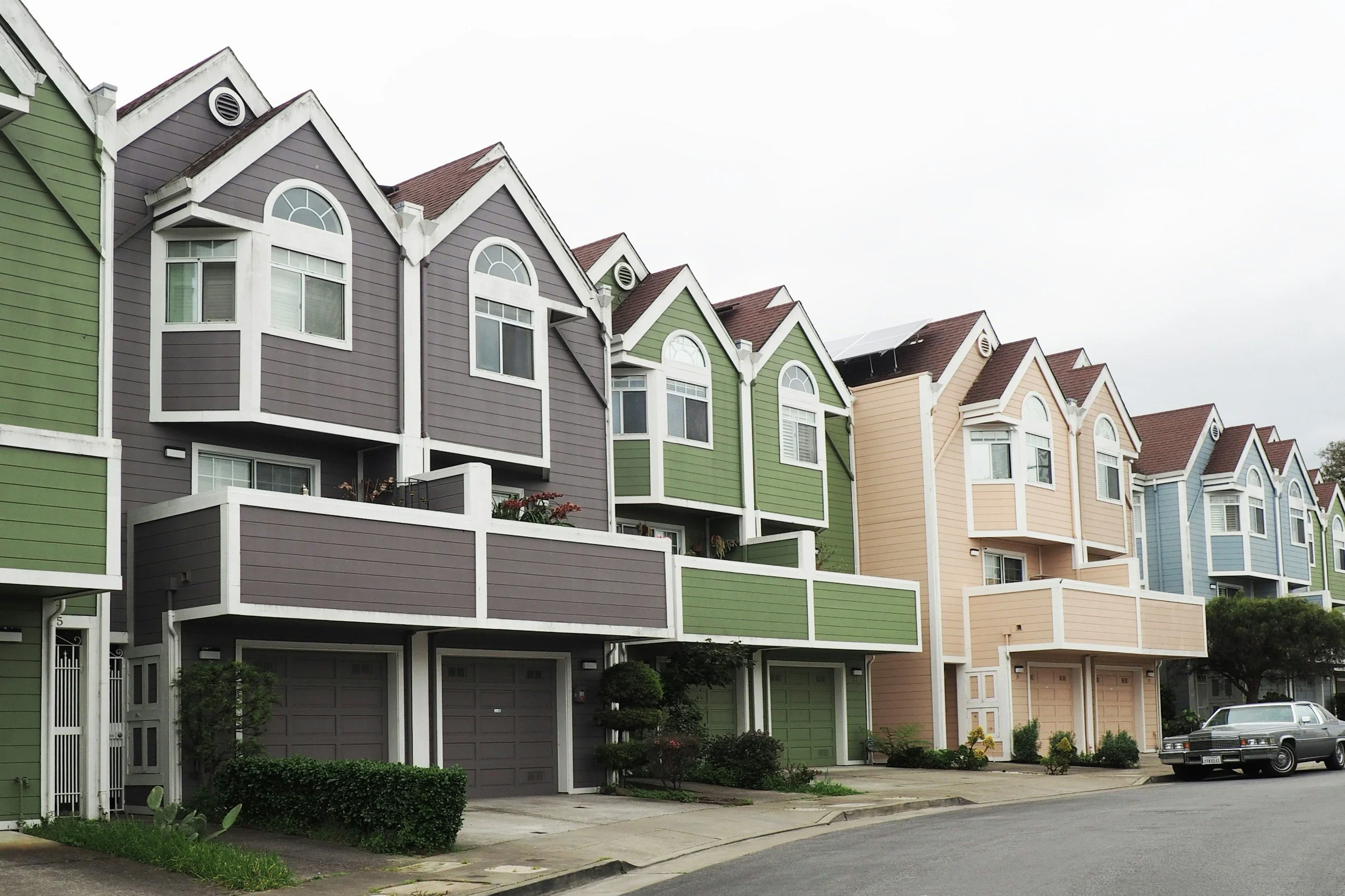Row of colorful multi-story townhouses with garages, small front gardens, and parked cars, under an overcast sky.