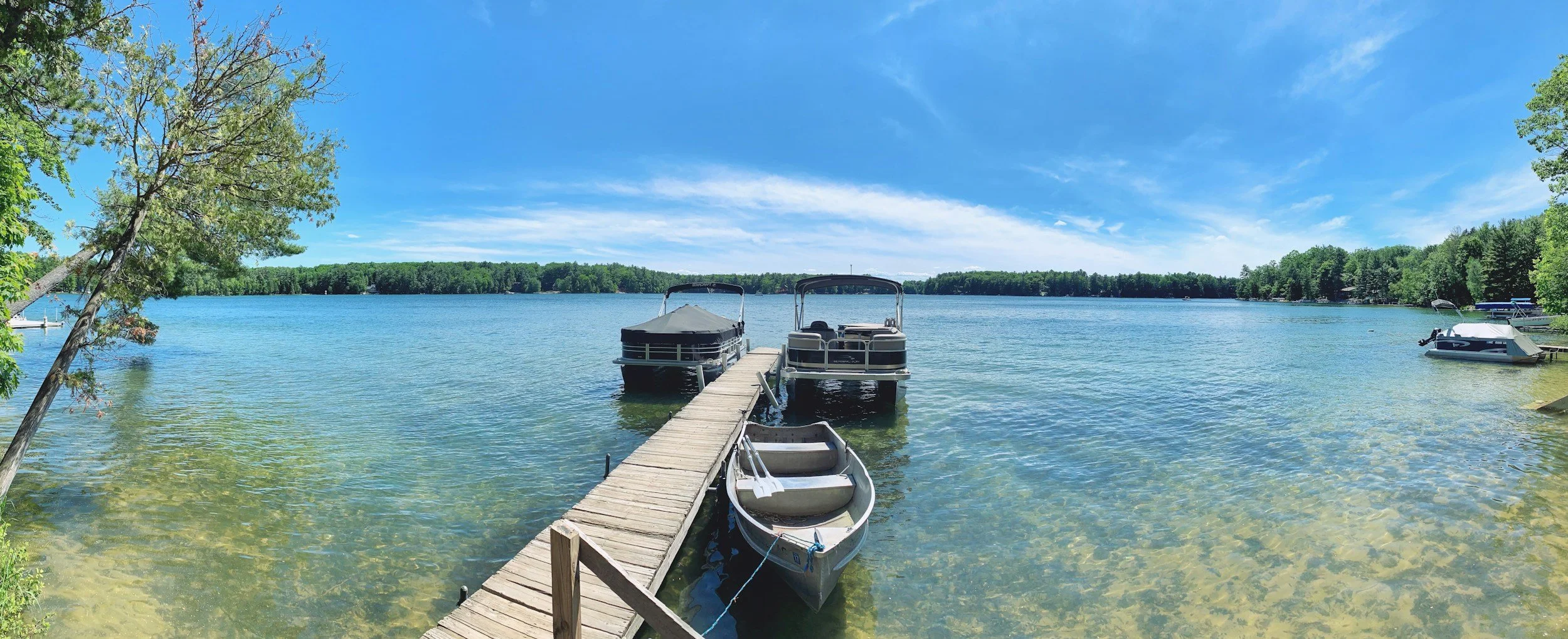 A wooden dock extending into a clear lake with boats tied to it, surrounded by green trees and a blue sky.