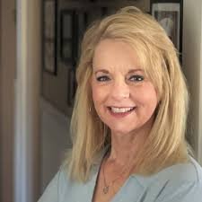 A smiling woman with blonde hair wearing a light-colored blazer standing indoors.