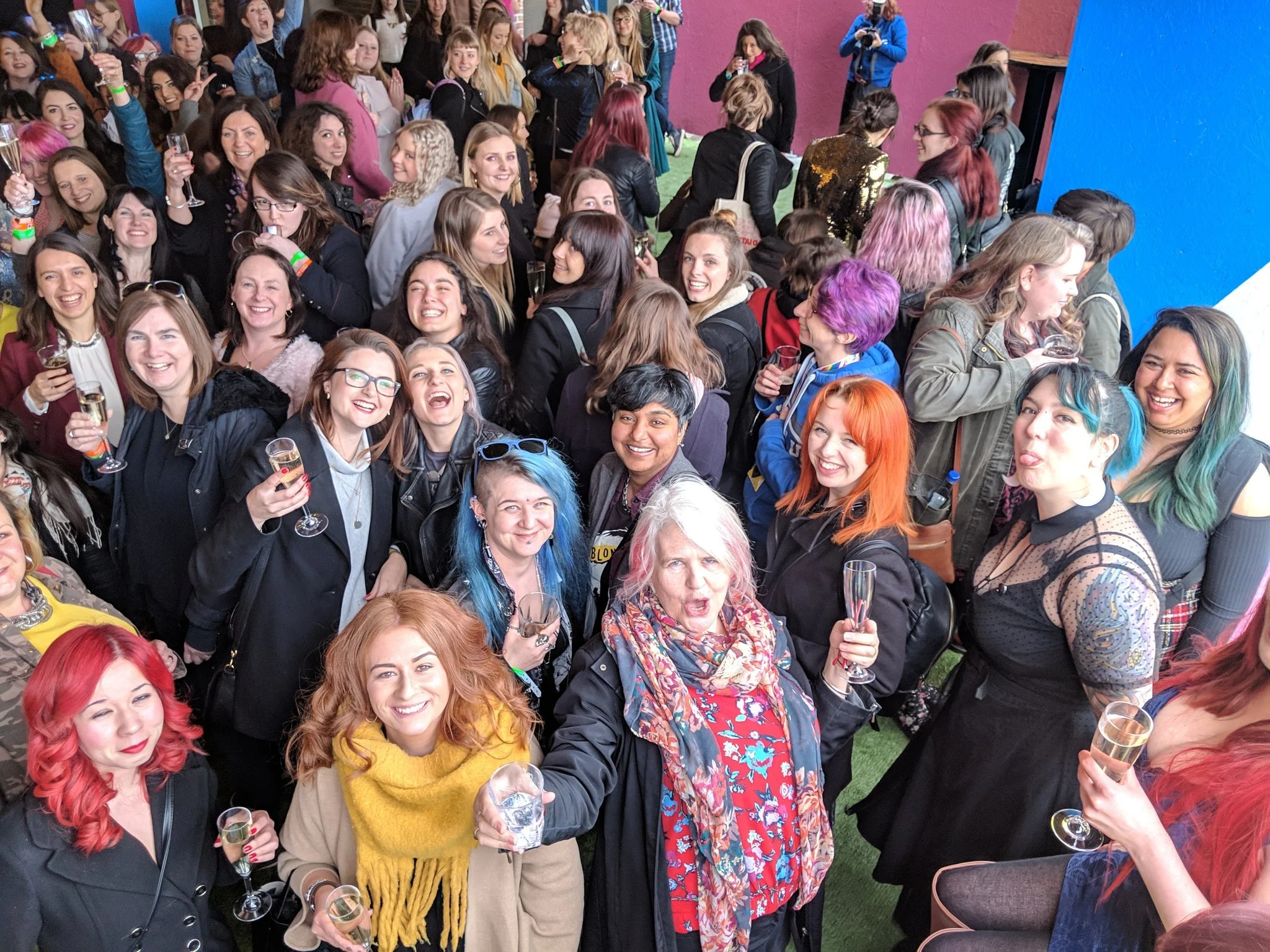 A large group of mostly women smiling, holding glasses of champagne at a social event or party, with colorful hair and diverse fashion styles.