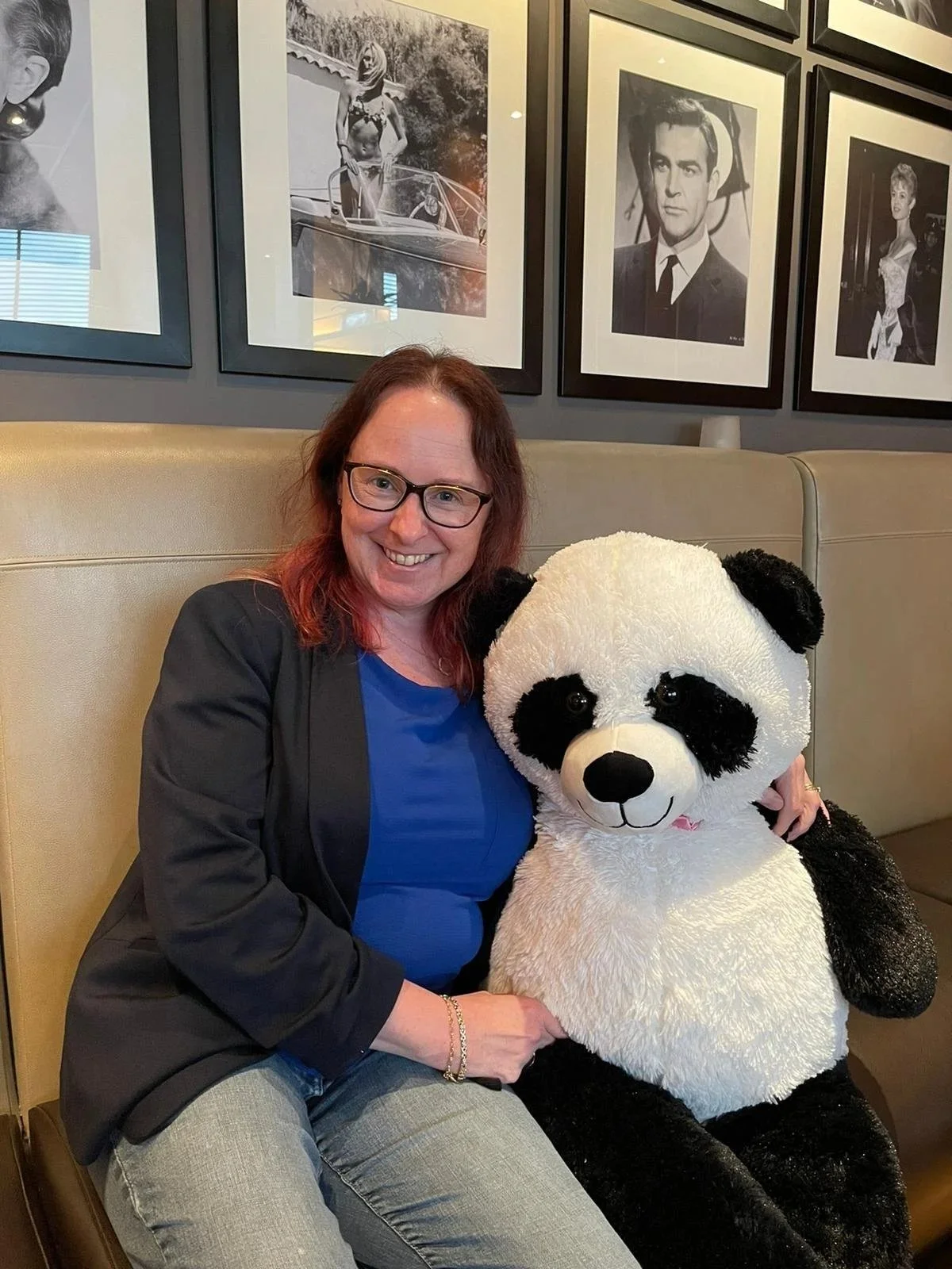 A woman with glasses smiling, sitting on a beige bench, holding a large stuffed panda bear, in a room with black and white photographs on the wall.