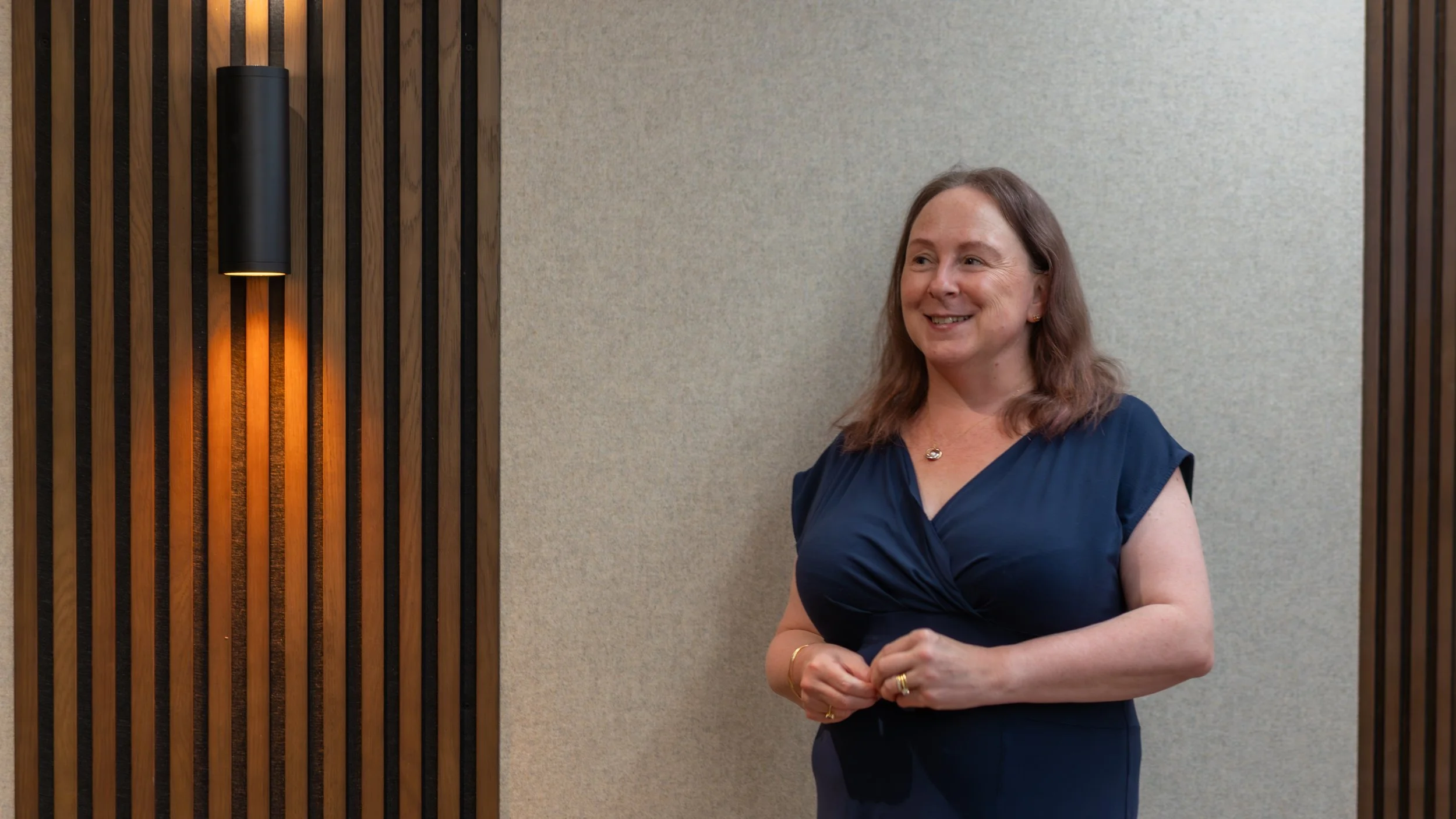 A woman with shoulder-length brown hair smiling, wearing a dark blue dress, standing in front of a wooden slat wall with a black wall sconce.