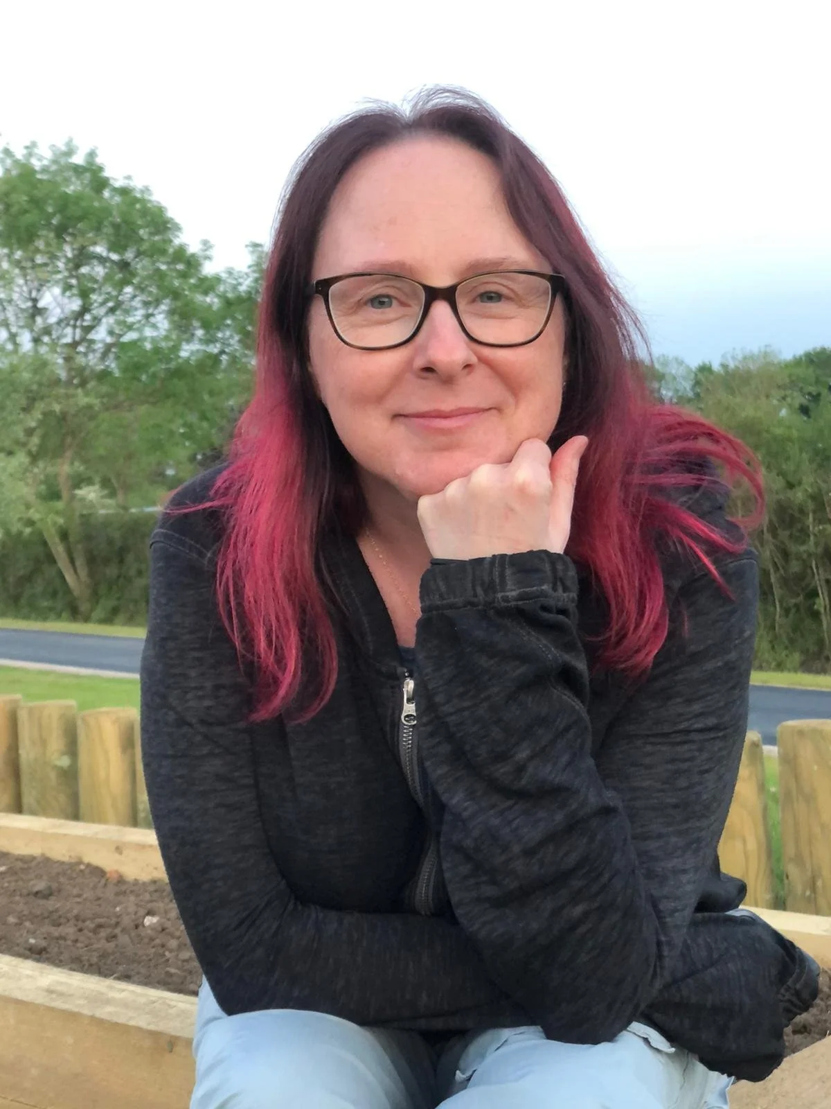A woman with glasses, long hair with dark and pink streaks, wearing a black zip-up jacket, sitting outdoors on a park bench with trees and a cloudy sky in the background.