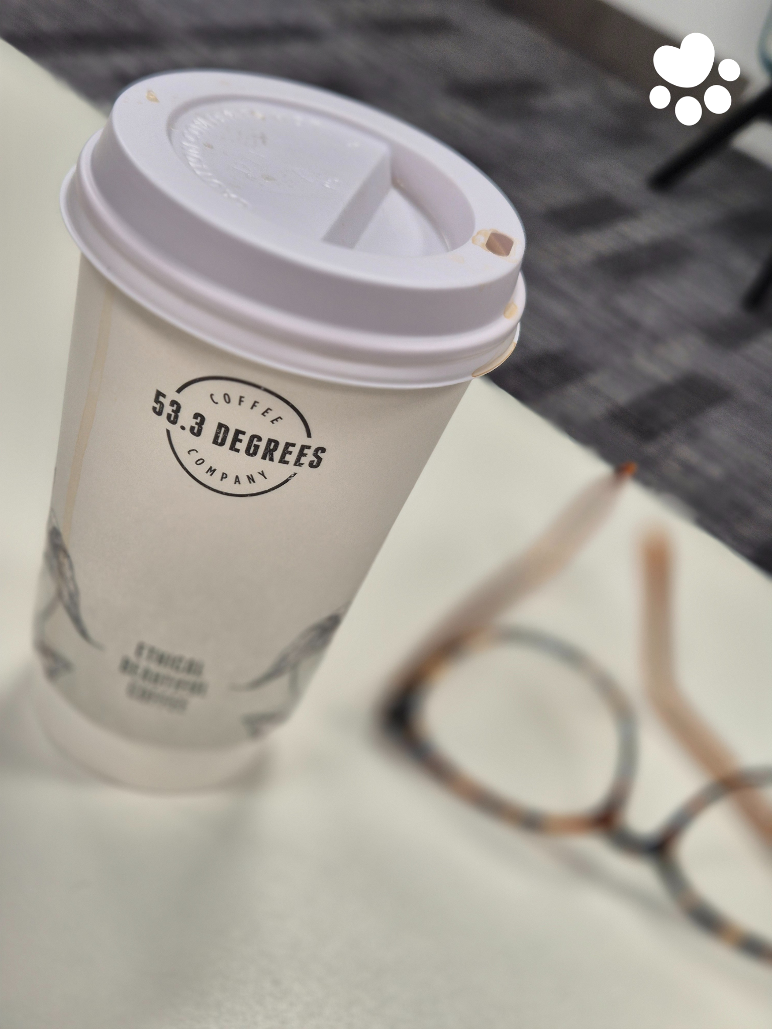 A disposable coffee cup with a plastic lid and a straw, placed on a white surface, with a pair of glasses in the background.