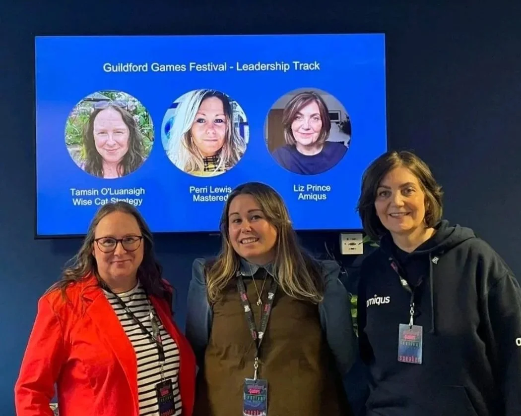 Three women standing in front of a screen displaying the Guildford Games Festival - Leadership Track, featuring photos and names of three speakers: Tamsin O'Luanaigh, Perri Lewis, and Liz Prince. The women are smiling and wearing festival badges.