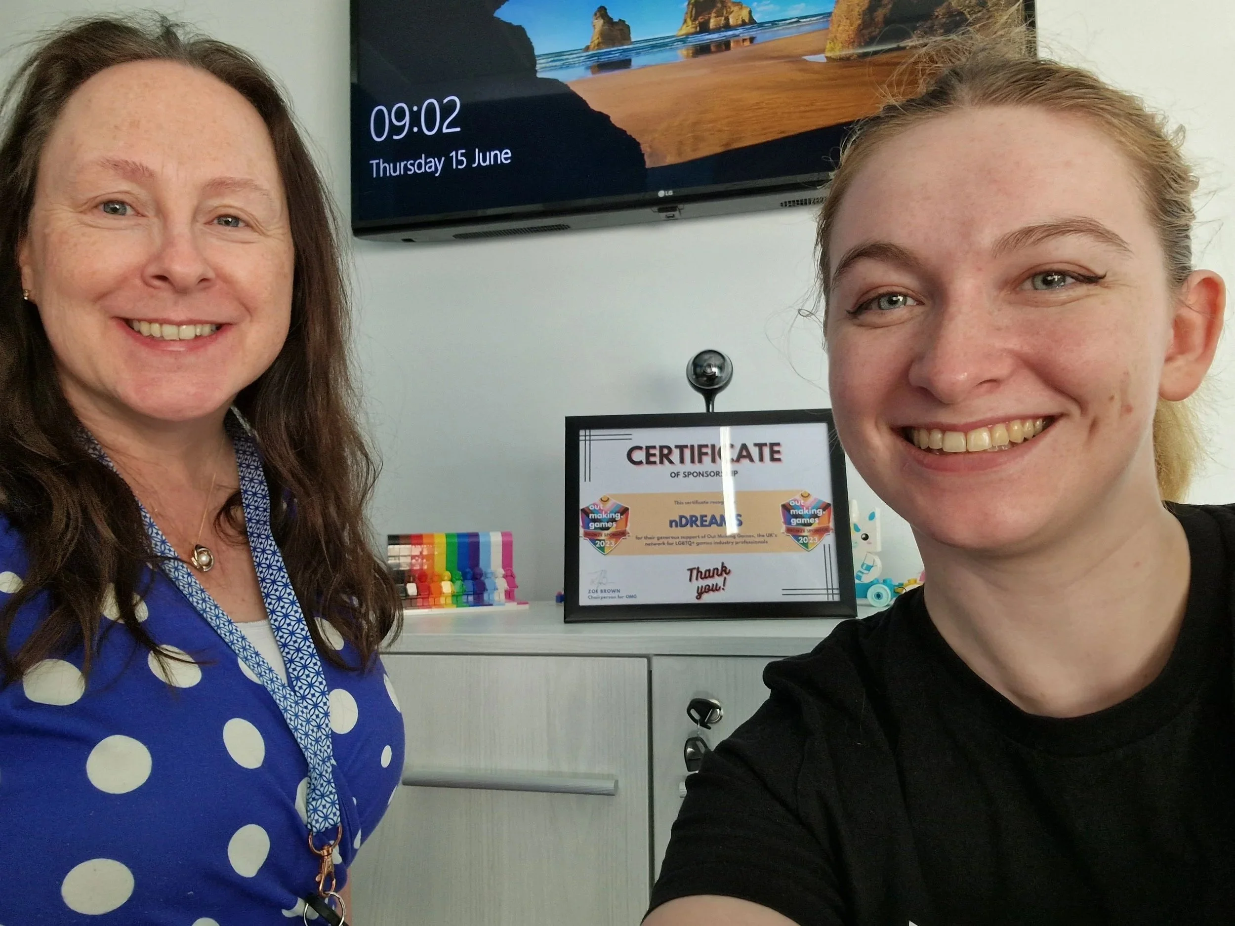 Two smiling women taking a selfie in an indoor setting, with a TV displaying the date and time and a certificate of sponsorship in the background.