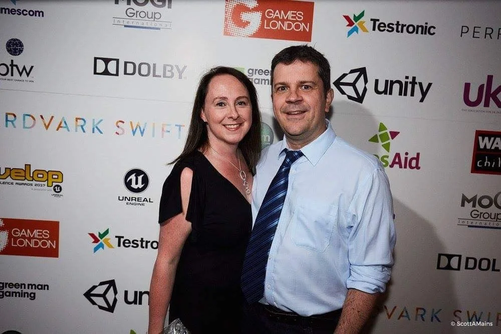A man and woman smiling and standing in front of a backdrop with multiple company logos at an event.